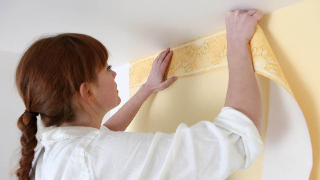 A woman putting up wallpaper border at the top of her ceiling. 