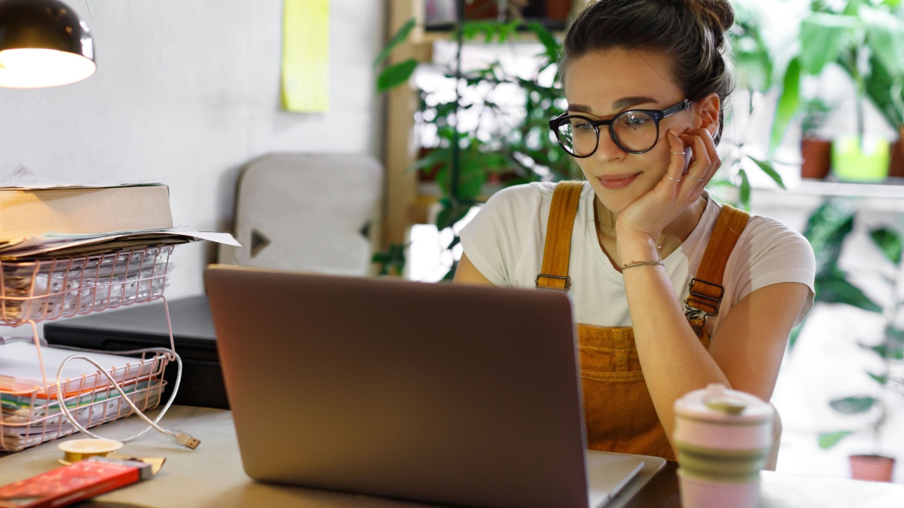 A young woman rests her head in her hand while working at her desk on her laptop.