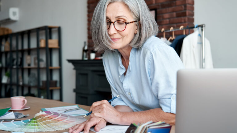 Middle aged stylish woman fashion designer drawing sketches in studio office. Mature old adult elegant grey-haired lady dressmaker small business owner creating new fashion design cloth in atelier.