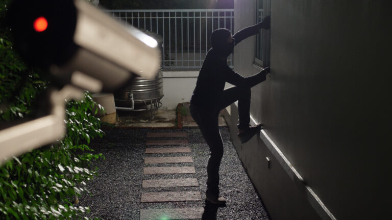 A disguised figure hoists into a first-floor window while filmed by a home security camera